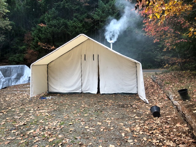A canvas prospector tent in the woods with smoke billowing from its chimney.
